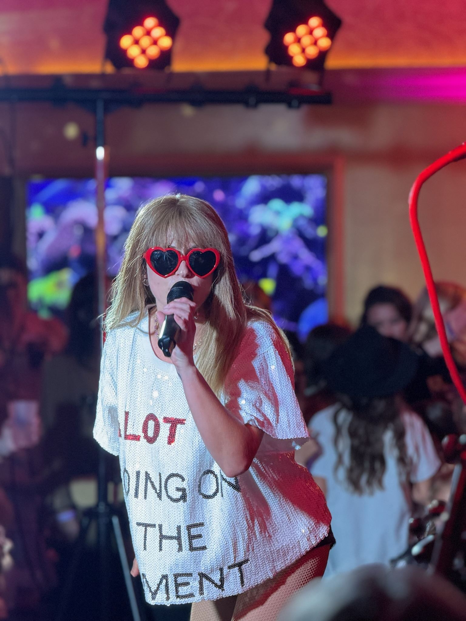 A woman in heart-shaped sunglasses and oversized white sequin shirt sings in front of a crowd.